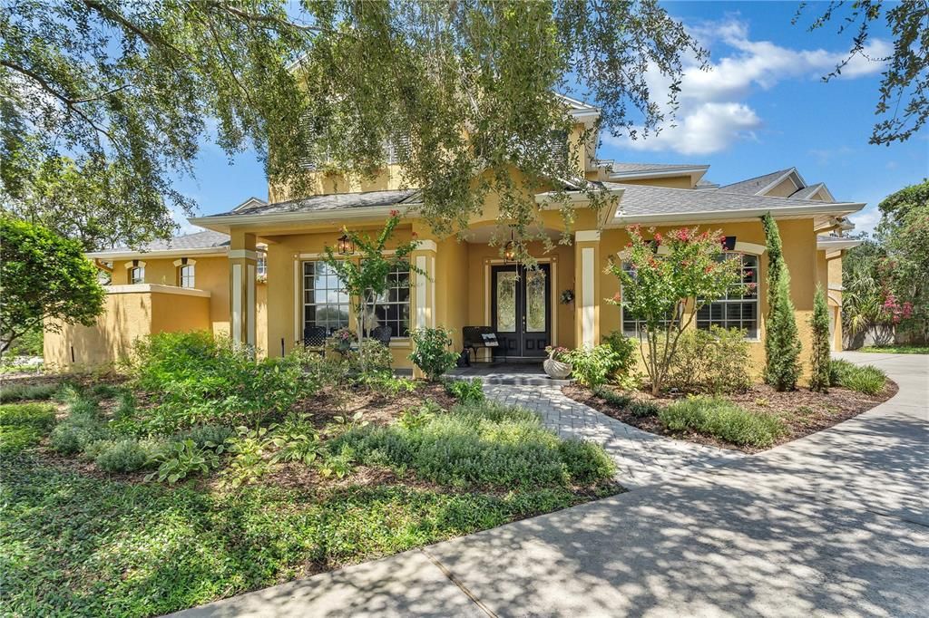 Two-storey stucco home with an arched entry, flowering shrubs and clipped hedges under a clear sky.