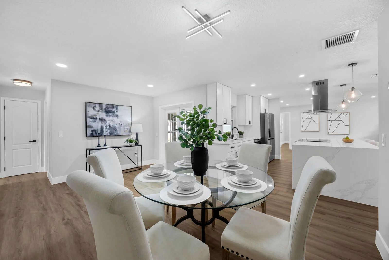 Open-plan dining area with a round glass table, upholstered chairs and a white kitchen beyond.